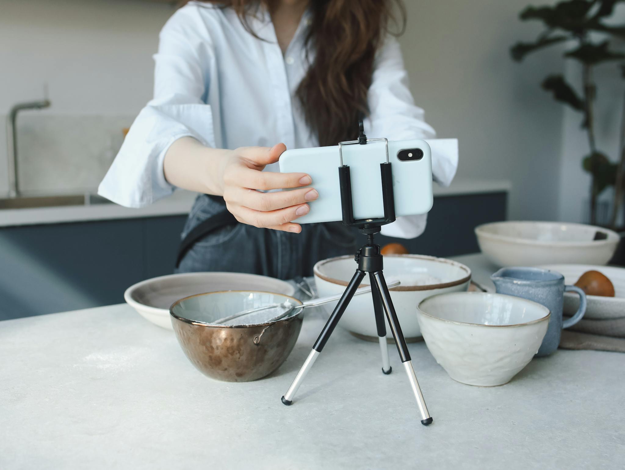 Woman using a smartphone on a tripod for vlogging in the kitchen with bowls.