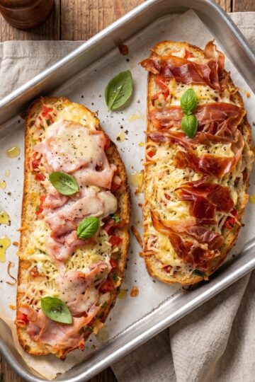 Savory baked bruschetta toasts with ham, olives, and melted cheese served on a baking tray.