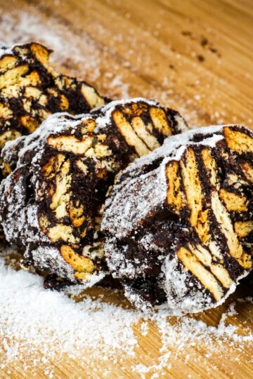 Sliced chocolate salami with cookie pieces and powdered sugar on a wooden board.