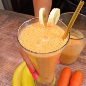 Glass of carrot banana juice decorated with banana slices and a gold straw, held by a hand with pink painted nails. Carrots and banana nearby.