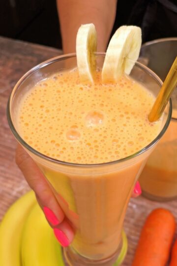 Glass of carrot banana juice decorated with banana slices and a gold straw, held by a hand with pink painted nails. Carrots and banana nearby.