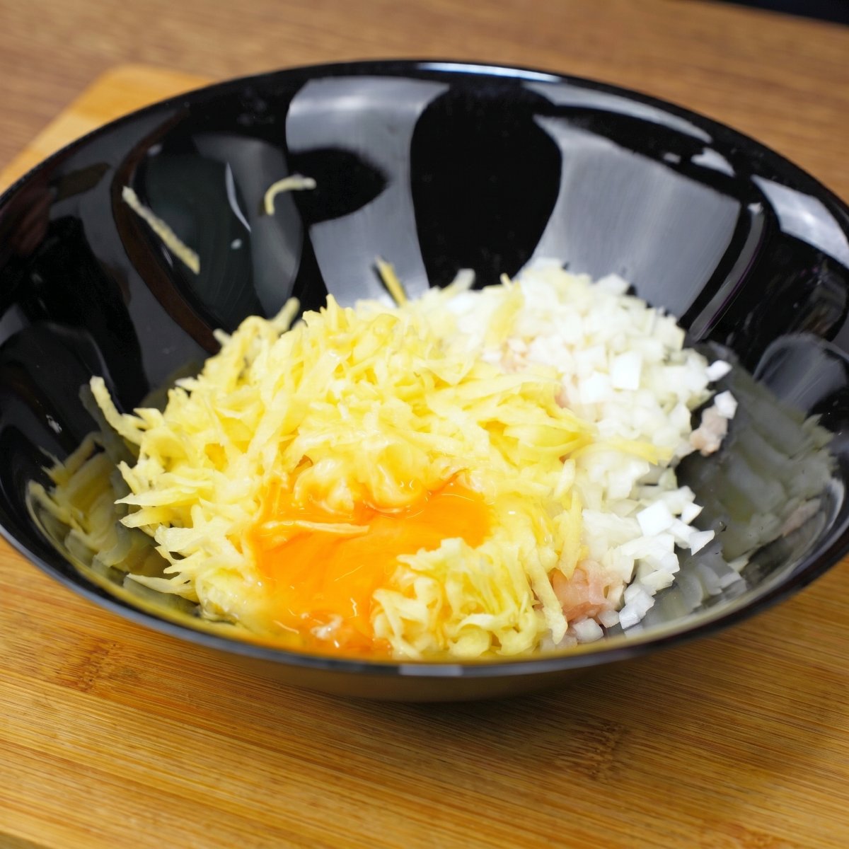 Ingredients for chicken and potato omelet in a black bowl: grated potato, egg yolk, chicken pieces, and onion.