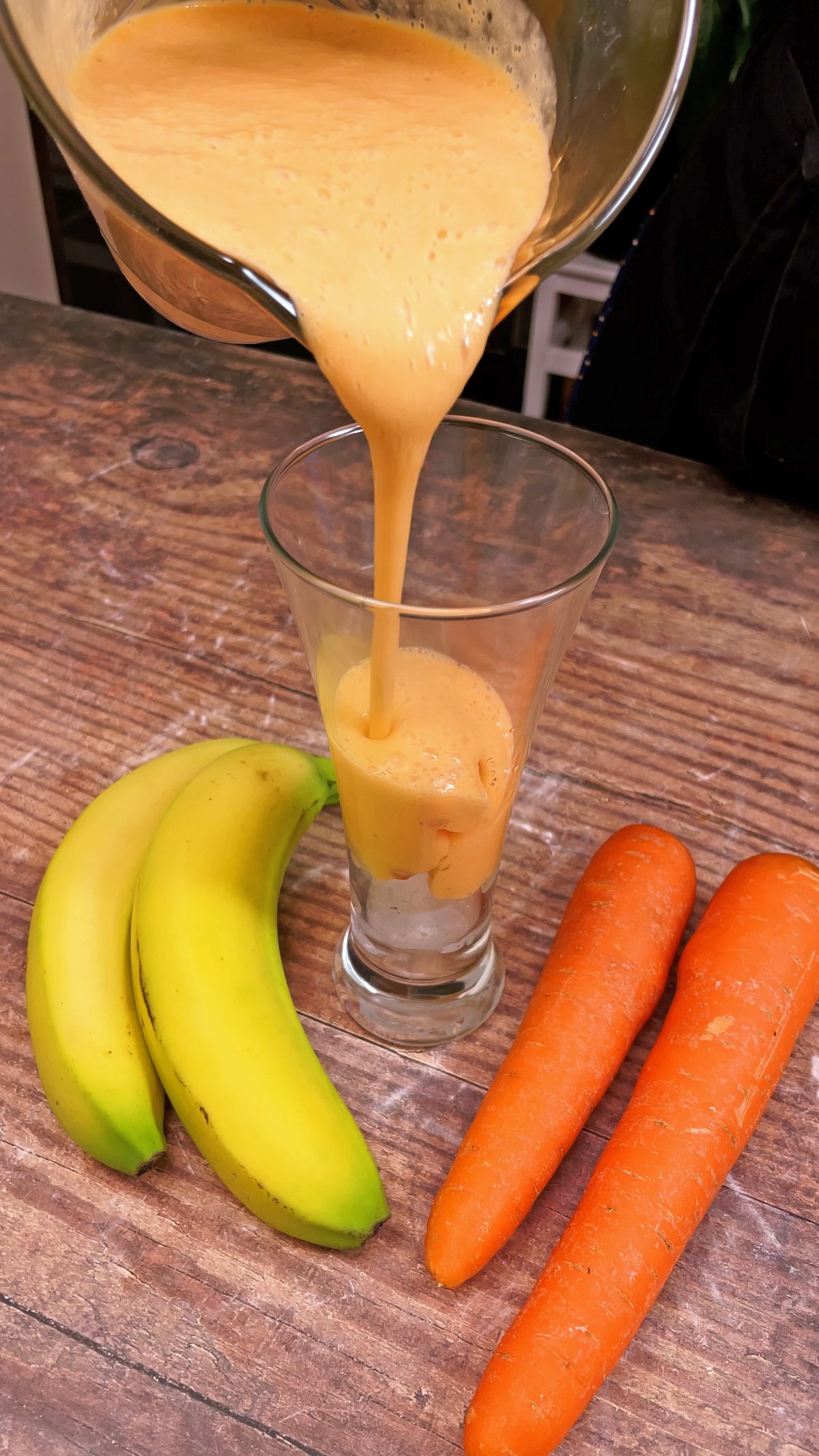 Karotten-Bananen-Saft wird in ein hohes Glas gegossen, daneben zwei Bananen und zwei Karotten auf einem Holztisch.